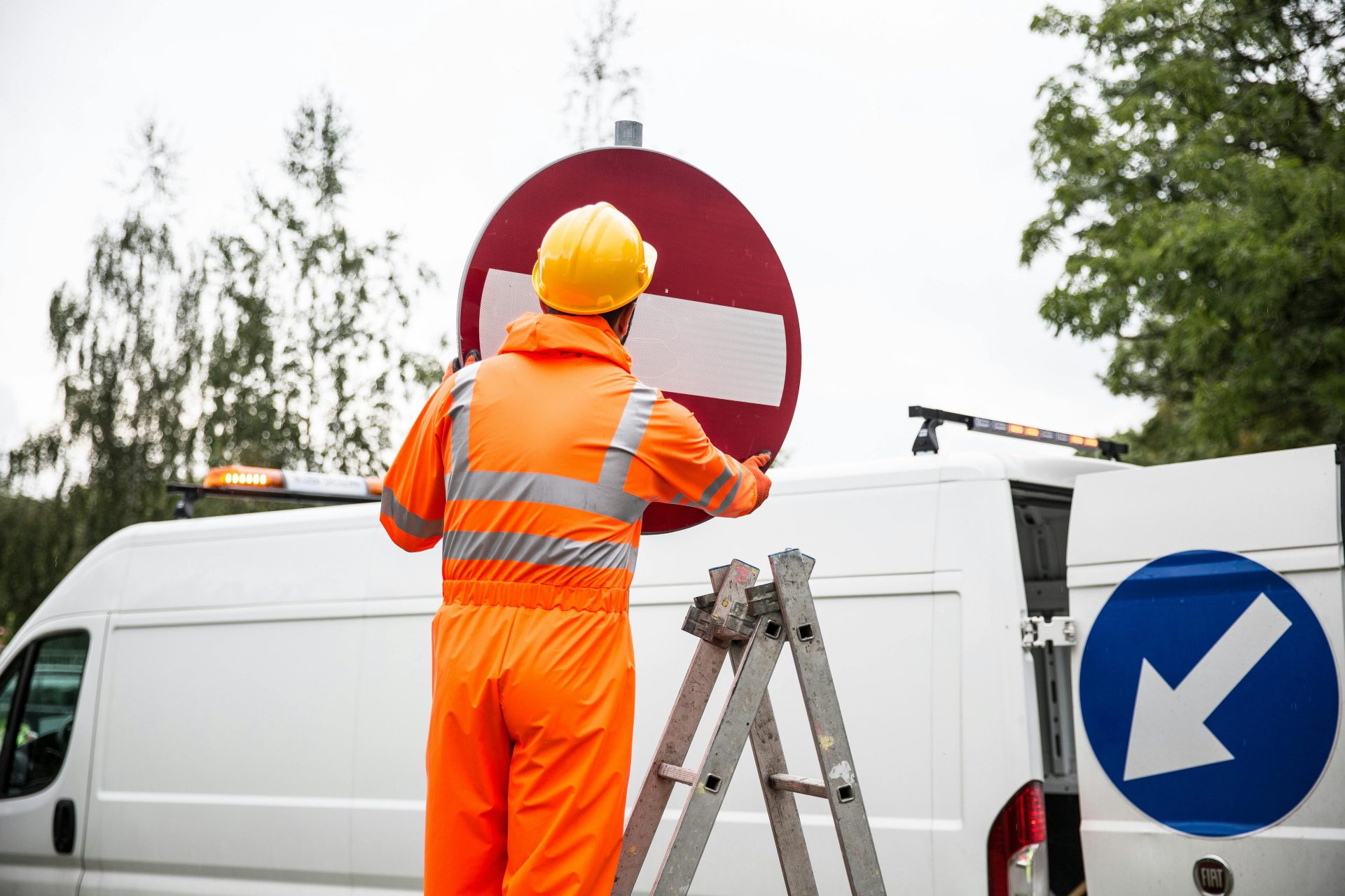 A road worker in orange uniform adjusting a traffic sign on a ladder by a van.